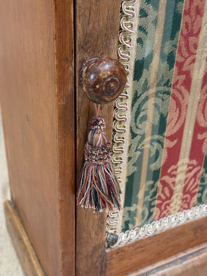 Wooden bedside cabinet with patterned fabric-inset door, braided gimp trim, tassel knob and carved egg-and-dart detailing. 