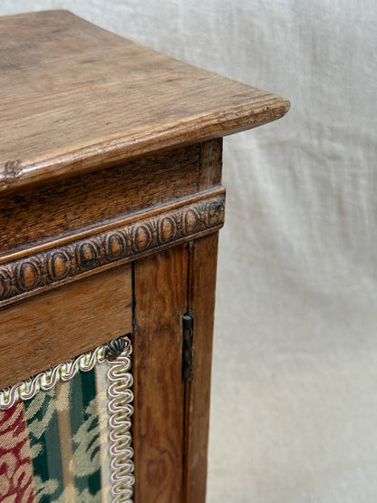 Wooden bedside cabinet with patterned fabric-inset door, braided gimp trim, tassel knob and carved egg-and-dart detailing. 