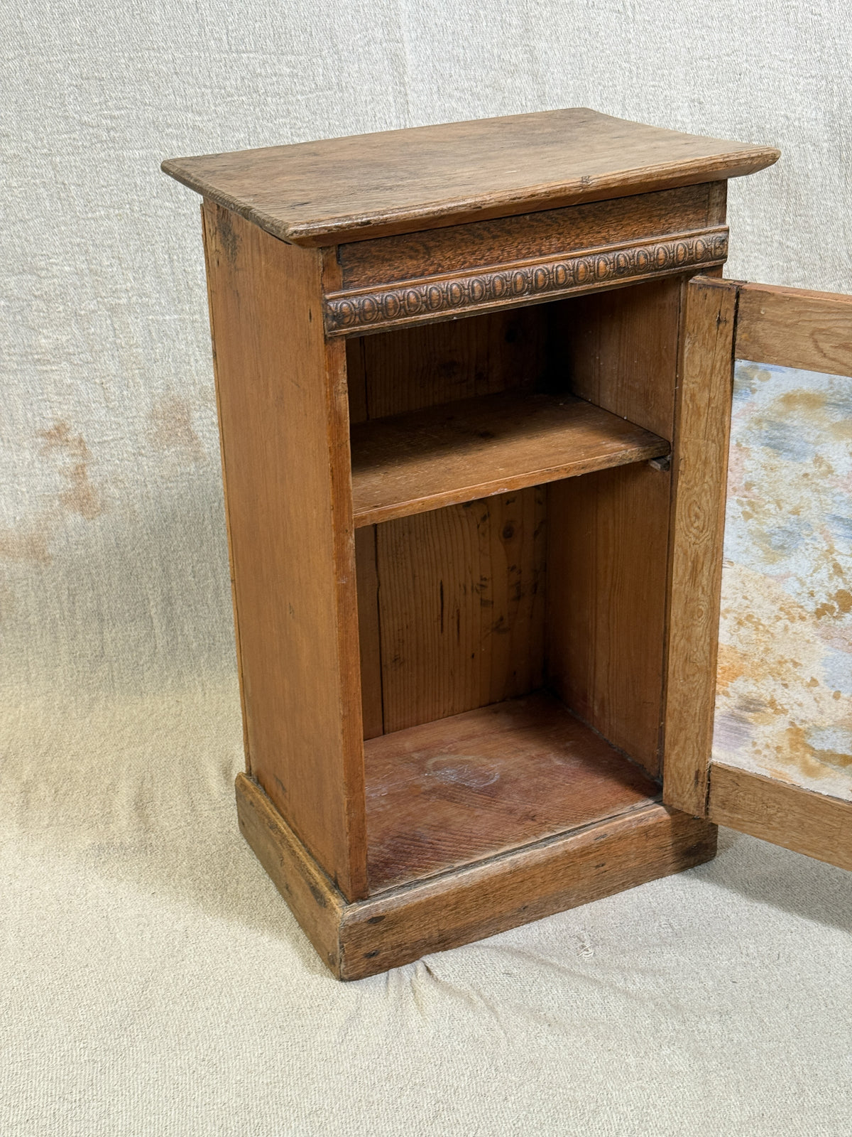 Wooden bedside cabinet with patterned fabric-inset door, braided gimp trim, tassel knob and carved egg-and-dart detailing. 
