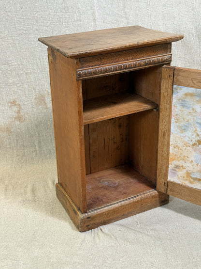 Wooden bedside cabinet with patterned fabric-inset door, braided gimp trim, tassel knob and carved egg-and-dart detailing. 