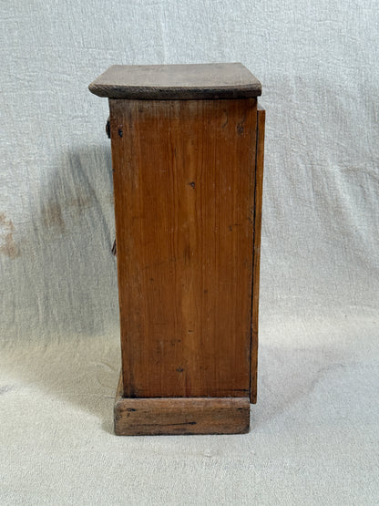 Wooden bedside cabinet with patterned fabric-inset door, braided gimp trim, tassel knob and carved egg-and-dart detailing. 
