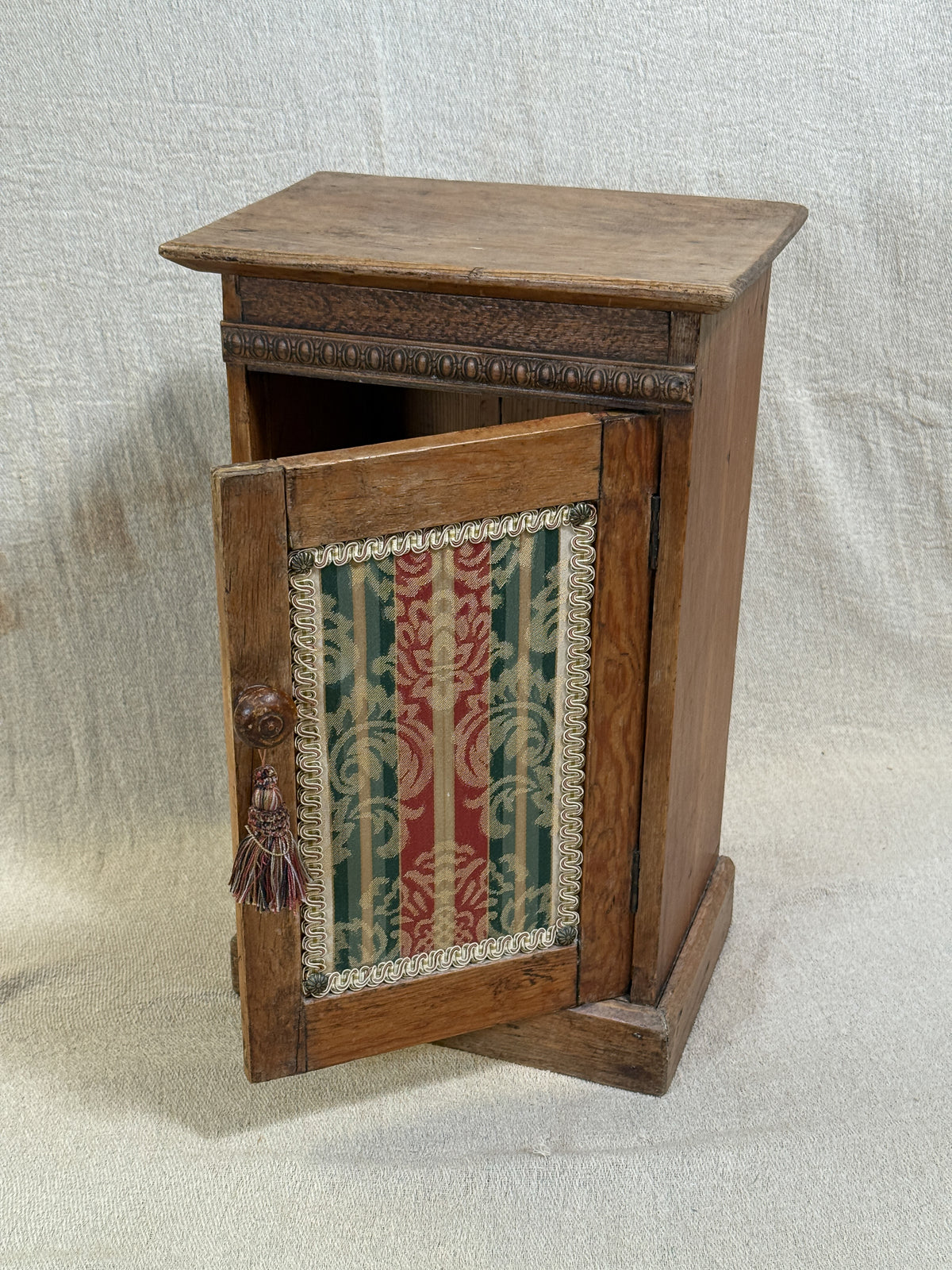 Wooden bedside cabinet with patterned fabric-inset door, braided gimp trim, tassel knob and carved egg-and-dart detailing. 