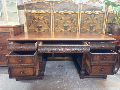 Vintage wooden desk with intricate carvings and multiple drawers in a room setting.