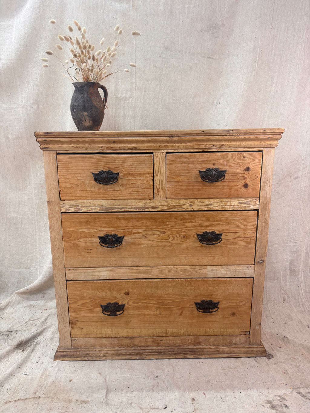 Wooden dresser with four drawers and a vase of dried flowers on top against a plain background