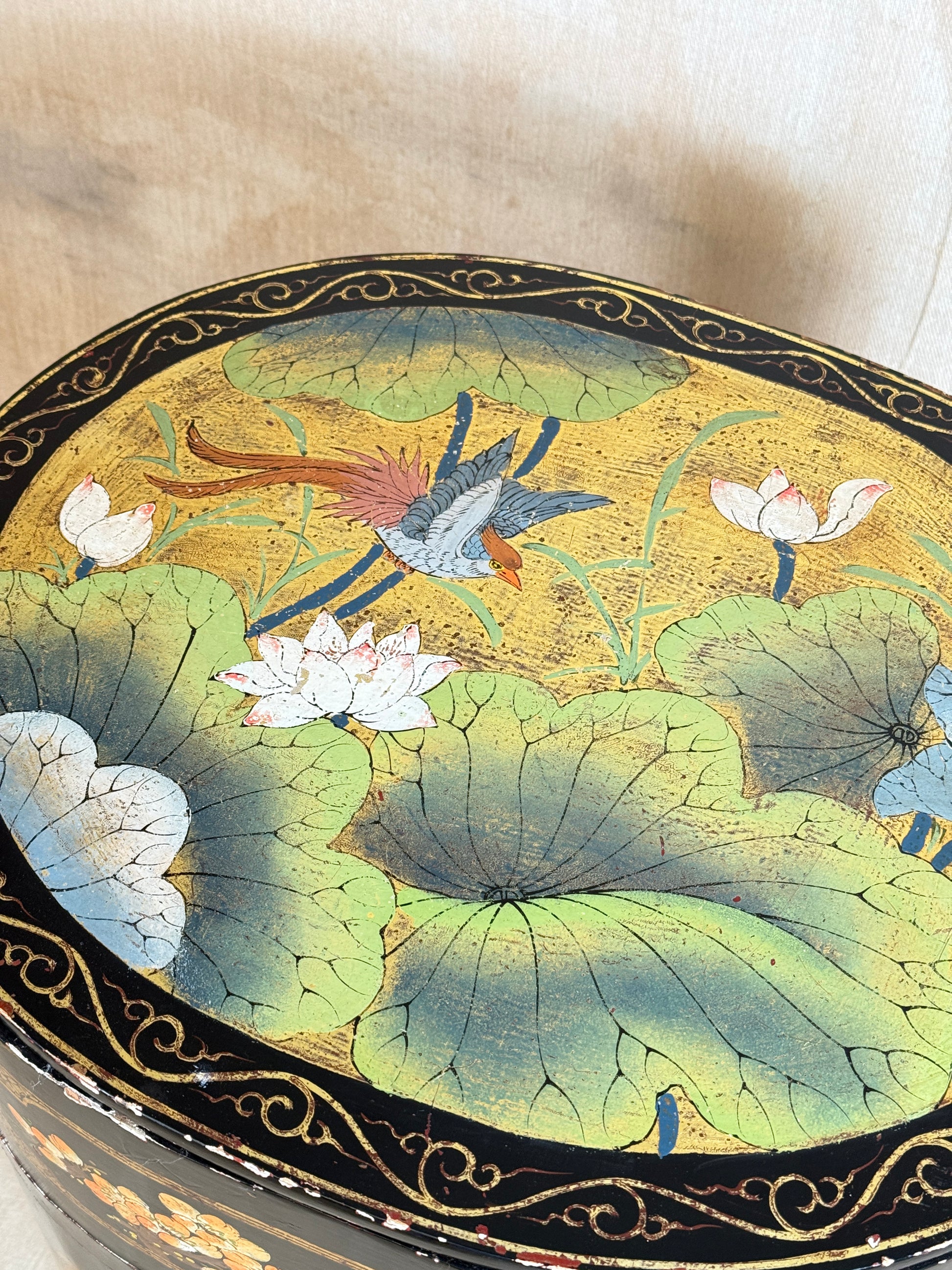 A close-up of a round decorative box lid featuring a hand-painted lotus pond scene with green lily pads, white lotus flowers, and birds on a gold background, bordered by a black and gold scroll pattern.