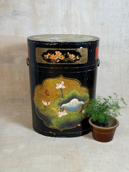 A vintage black lacquered cylindrical storage box with hand-painted lotus pond and floral motifs, displayed next to a small potted green plant on a neutral textured beige background.