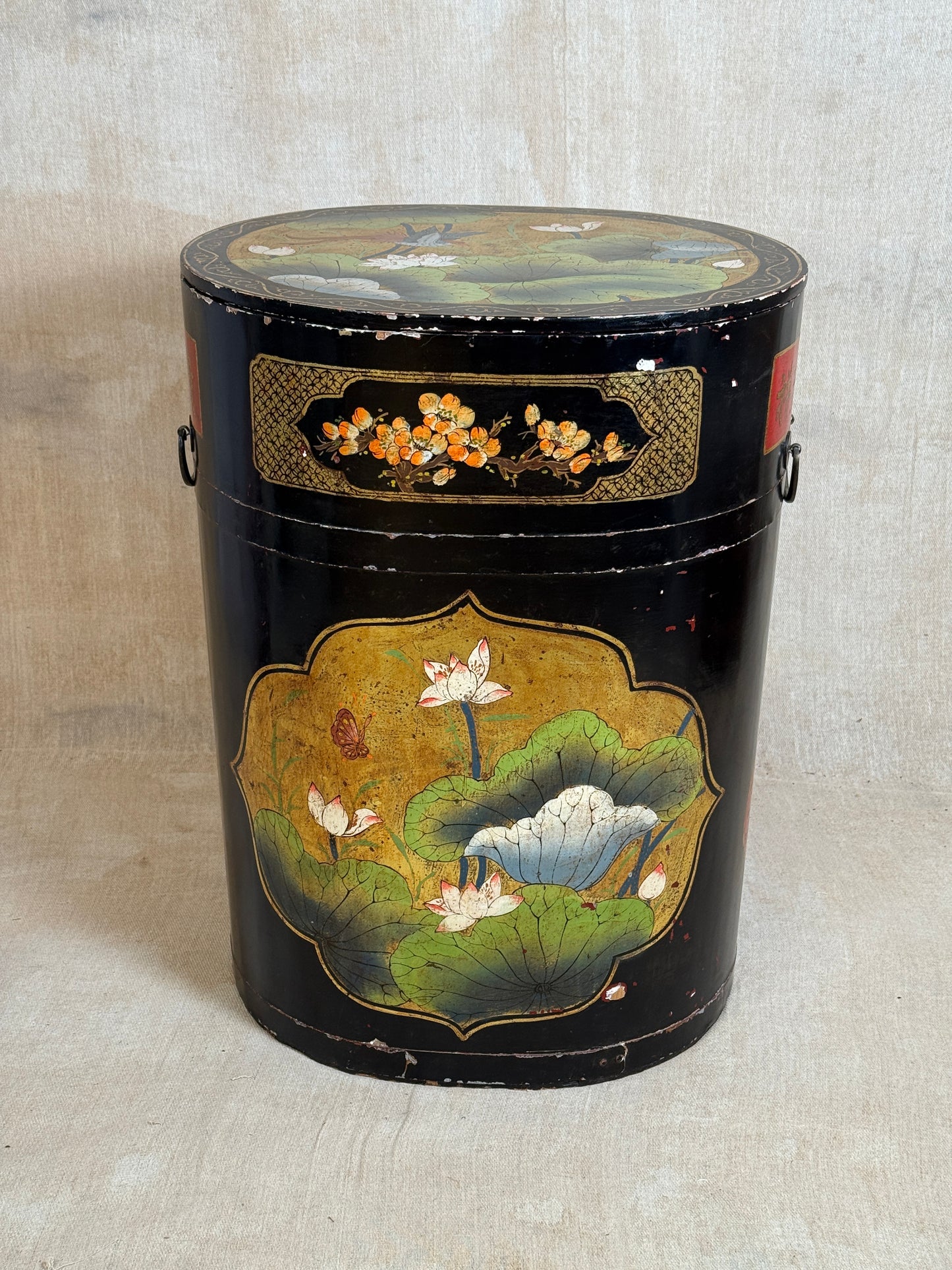 A vintage black lacquered cylindrical storage box decorated with hand-painted lotus pond scenes, featuring white lotus flowers, green leaves, and gold accents, set against a neutral textured backdrop.