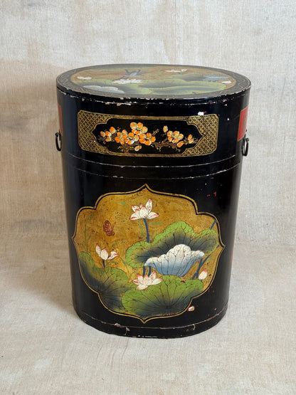 A vintage black lacquered cylindrical storage box decorated with hand-painted lotus pond scenes, featuring white lotus flowers, green leaves, and gold accents, set against a neutral textured backdrop.