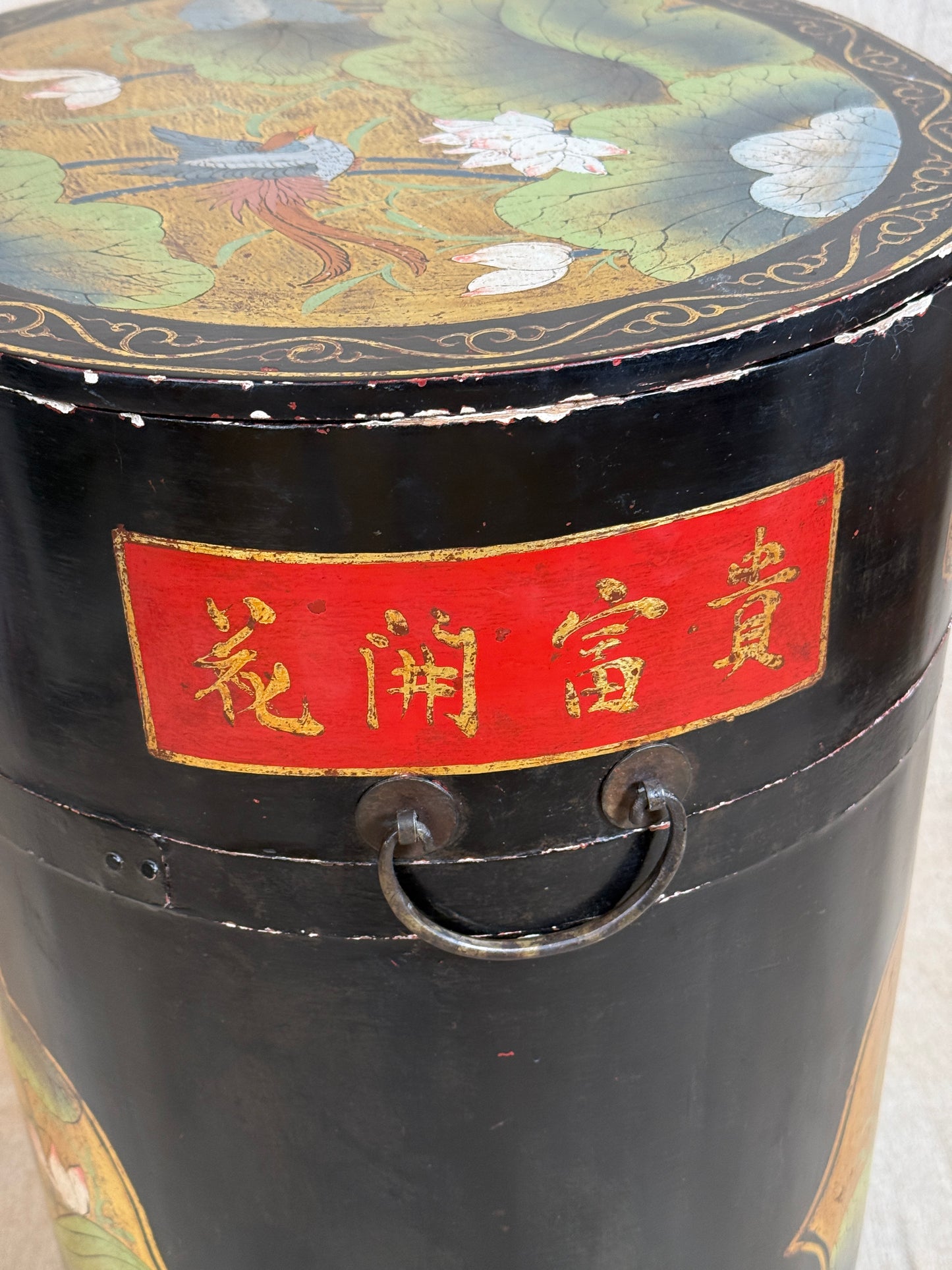 A close-up of a black cylindrical decorative container or canister with a red label featuring gold Chinese characters and a metal ring handle. The top lid is decorated with a painted lotus scene in green, gold, white, and blue tones.