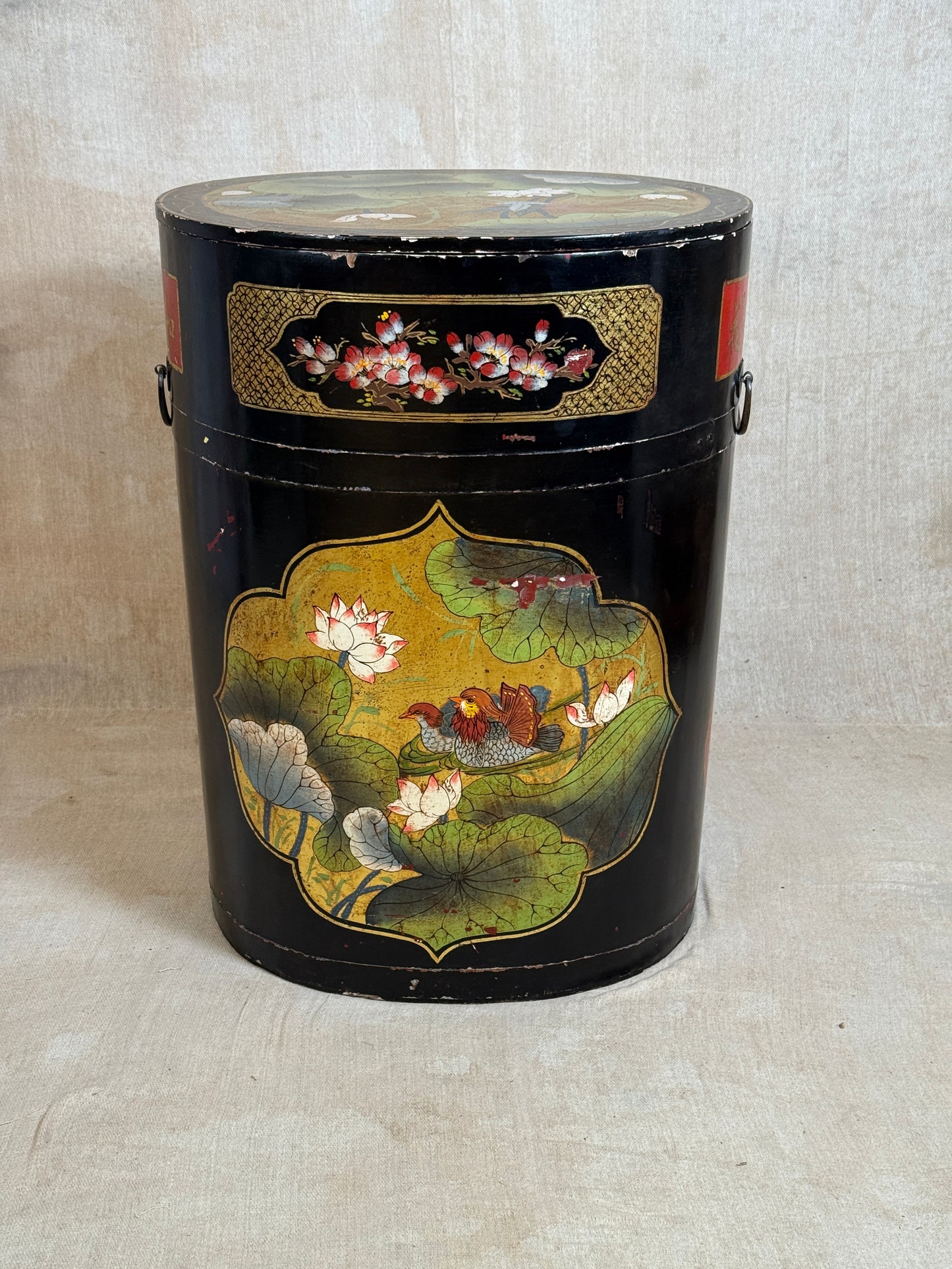 A vintage black metal canister with hand-painted lotus pond and fish motifs, featuring gold decorative panels and side handles, set against a neutral beige fabric backdrop.