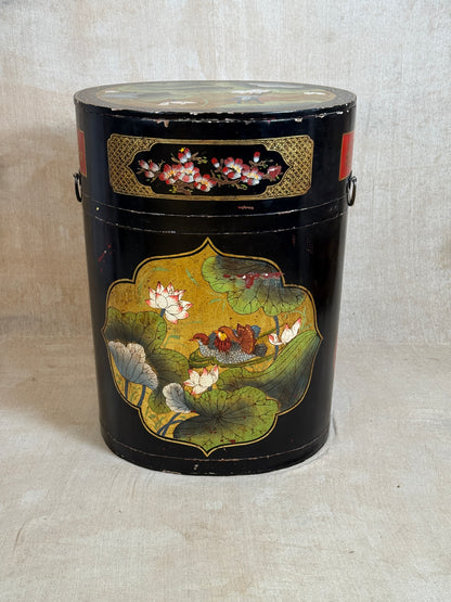 A vintage black metal canister with hand-painted lotus pond and fish motifs, featuring gold decorative panels and side handles, set against a neutral beige fabric backdrop.