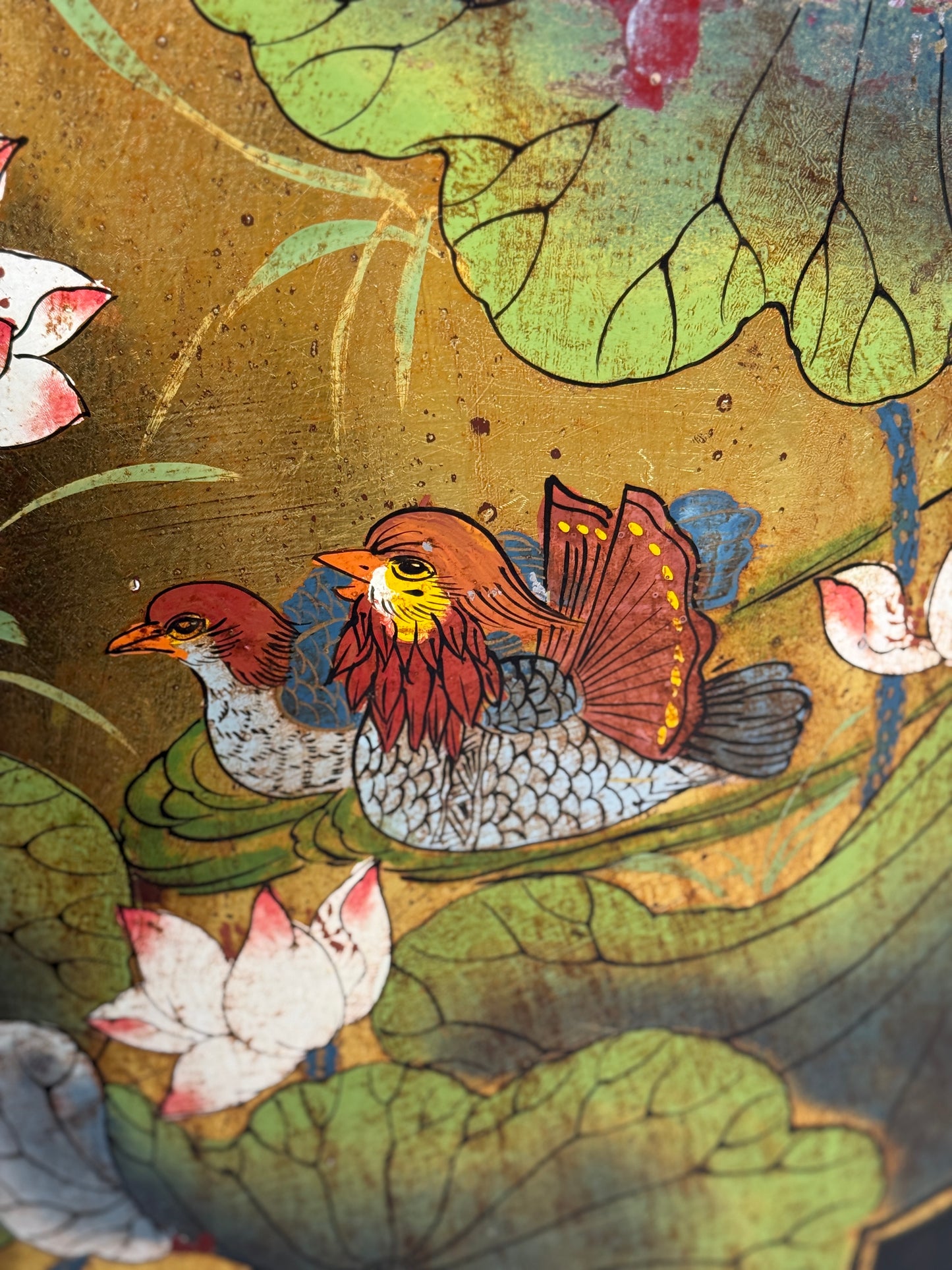 Close-up of a decorative artwork showing two birds among large green lily pads and white flowers on a textured gold background.