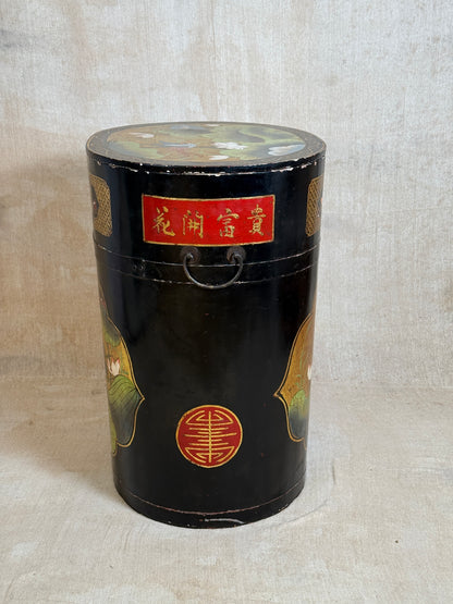 A vintage black lacquered cylindrical storage box with hand-painted gold and multicolor details, featuring a red label with gold Chinese characters and a metal handle, set against a neutral beige fabric background.