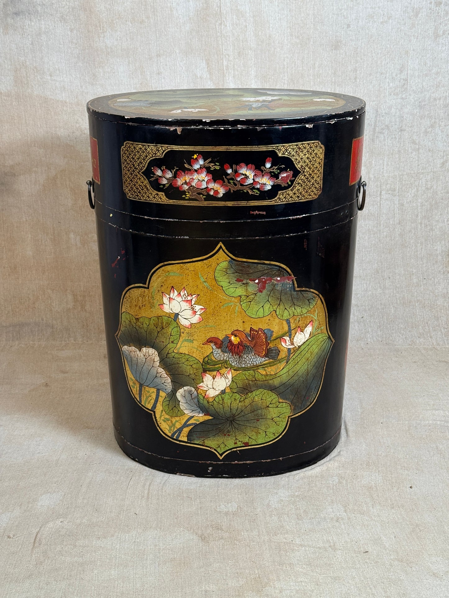 A vintage black cylindrical storage canister decorated with hand-painted lotus flowers and green leaves, featuring gold accents and metal side handles, set against a neutral beige fabric backdrop.