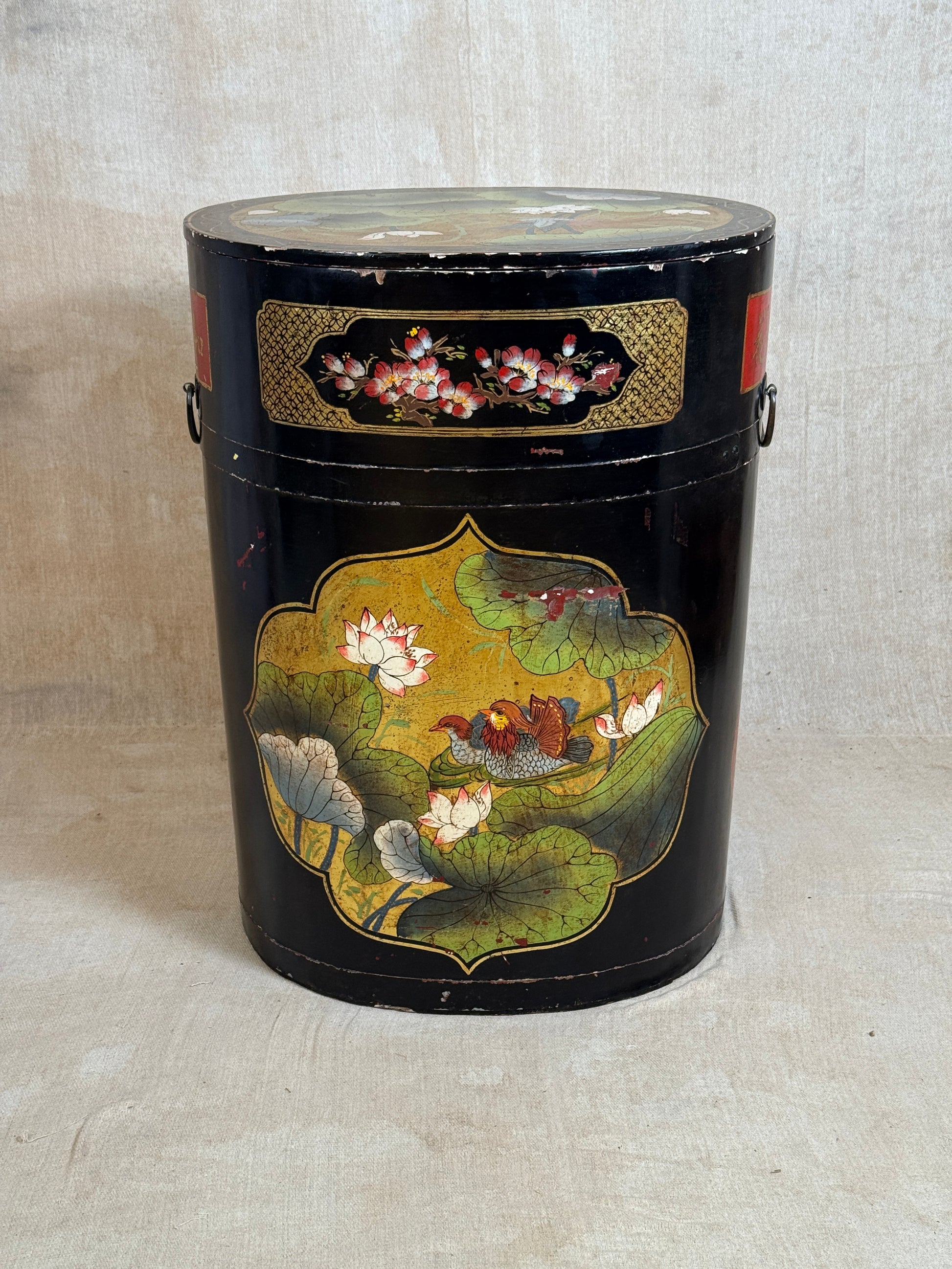 A vintage black cylindrical storage canister decorated with hand-painted lotus flowers and green leaves, featuring gold accents and metal side handles, set against a neutral beige fabric backdrop.