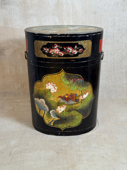 A vintage black cylindrical storage canister decorated with hand-painted lotus flowers and green leaves, featuring gold accents and metal side handles, set against a neutral beige fabric backdrop.