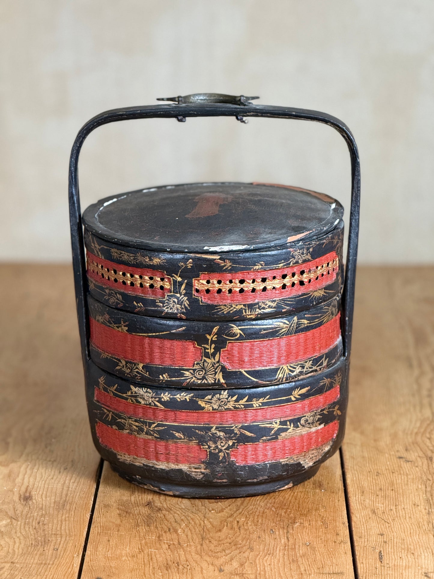 A vintage round wooden bento box with a black metal handle, featuring red and gold painted stripes and bamboo motifs, set on a wooden table against a neutral beige background.