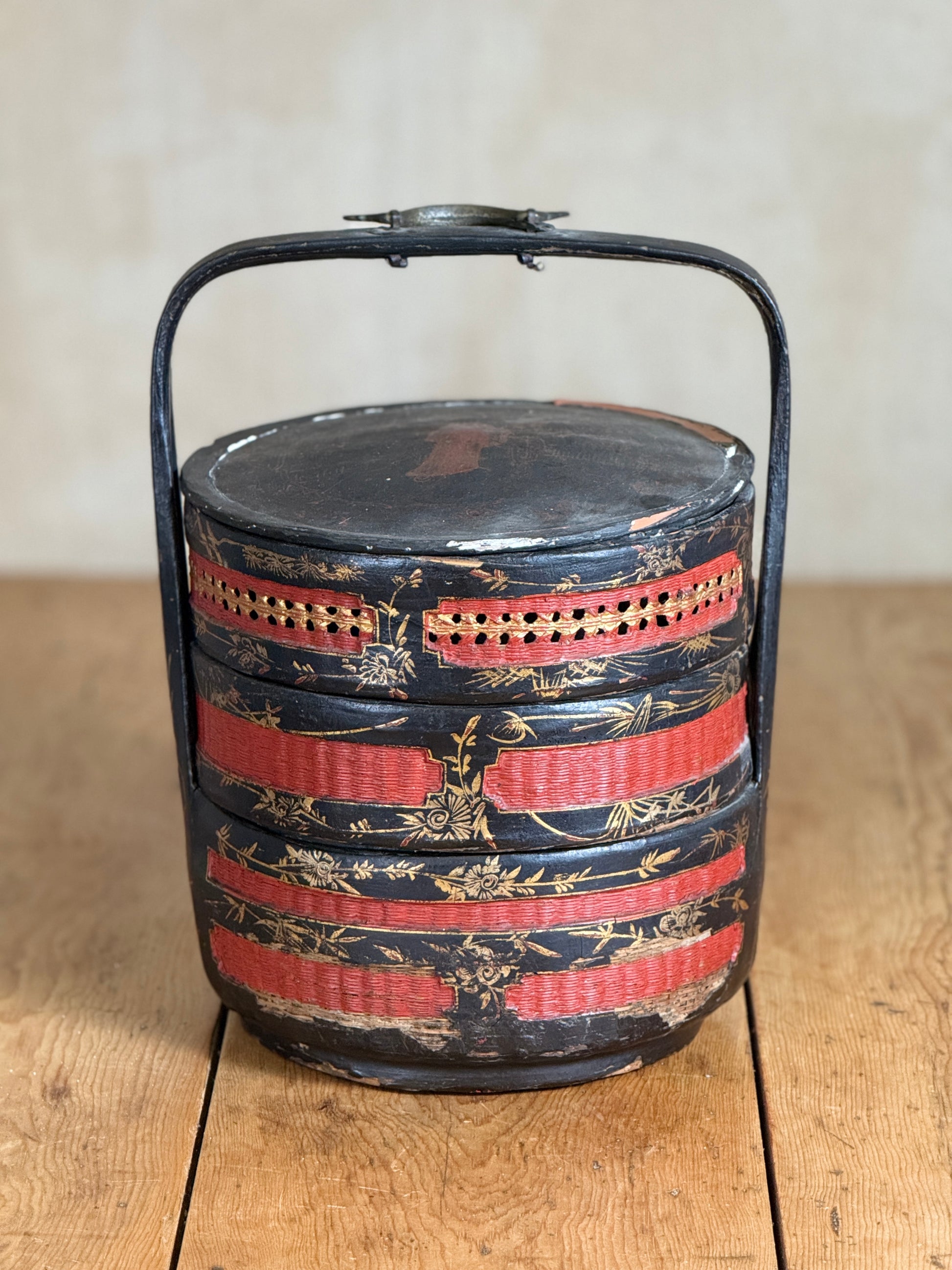 A vintage round wooden bento box with a black metal handle, featuring red and gold painted stripes and bamboo motifs, set on a wooden table against a neutral beige background.