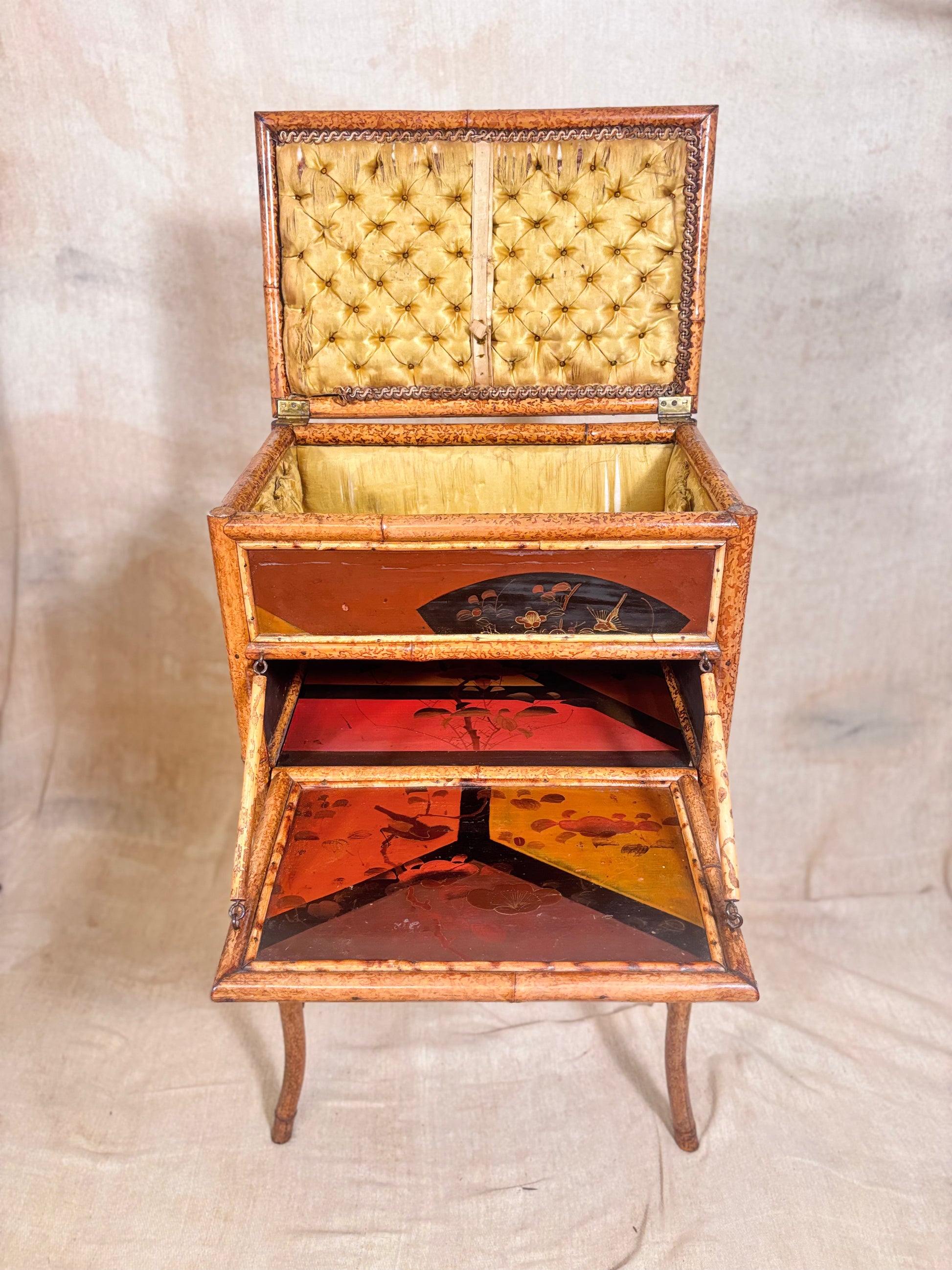 An antique folding bamboo chest or cabinet with an open top and lower drawer, featuring tufted yellow lining, red and gold painted interior panels, and brass hardware, photographed against a beige fabric backdrop.