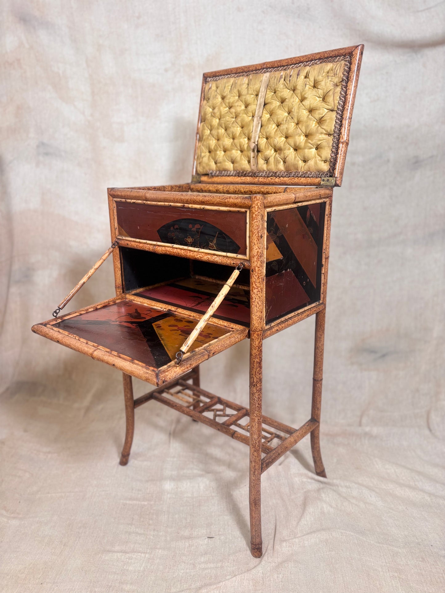 An antique folding sewing desk with a raised hinged top, upholstered in tufted yellow fabric, and a brown wooden frame with dark reddish-brown paneling. The desk is shown open, displaying its interior and lower writing surface, set against a neutral draped fabric backdrop.