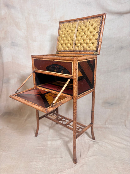An antique folding sewing desk with a raised hinged top, upholstered in tufted yellow fabric, and a brown wooden frame with dark reddish-brown paneling. The desk is shown open, displaying its interior and lower writing surface, set against a neutral draped fabric backdrop.