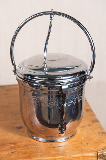 A vintage polished silver metal ice bucket with a carrying handle, shown on a wooden surface against a neutral beige background.