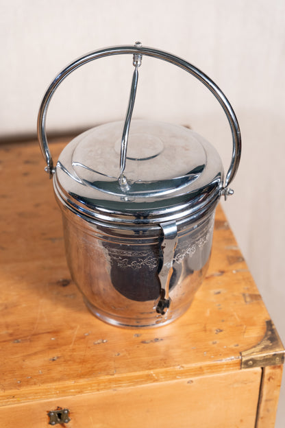 A vintage polished metal ice bucket with a carrying handle, resting on a rustic wooden chest against a neutral wall.
