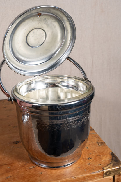 A vintage polished metal ice bucket with its lid propped open, resting on a rustic wooden surface against a neutral beige wall.
