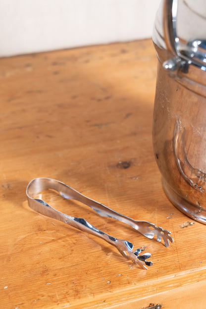 A pair of polished stainless steel ice tongs resting on a warm wooden surface, with a large silver metal container partially visible in the background.