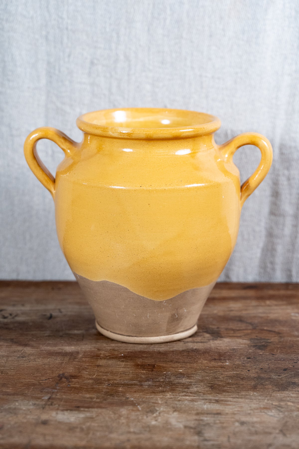 A handmade ceramic vase with a glossy yellow glaze on the upper half and a natural, unglazed clay base on the lower half, featuring two loop handles, set on a rustic wooden surface against a gray fabric backdrop.