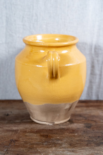 A rustic ceramic jar with a glossy mustard yellow glaze and a natural unglazed clay base, featuring a single loop handle, set on a wooden surface against a gray fabric backdrop.