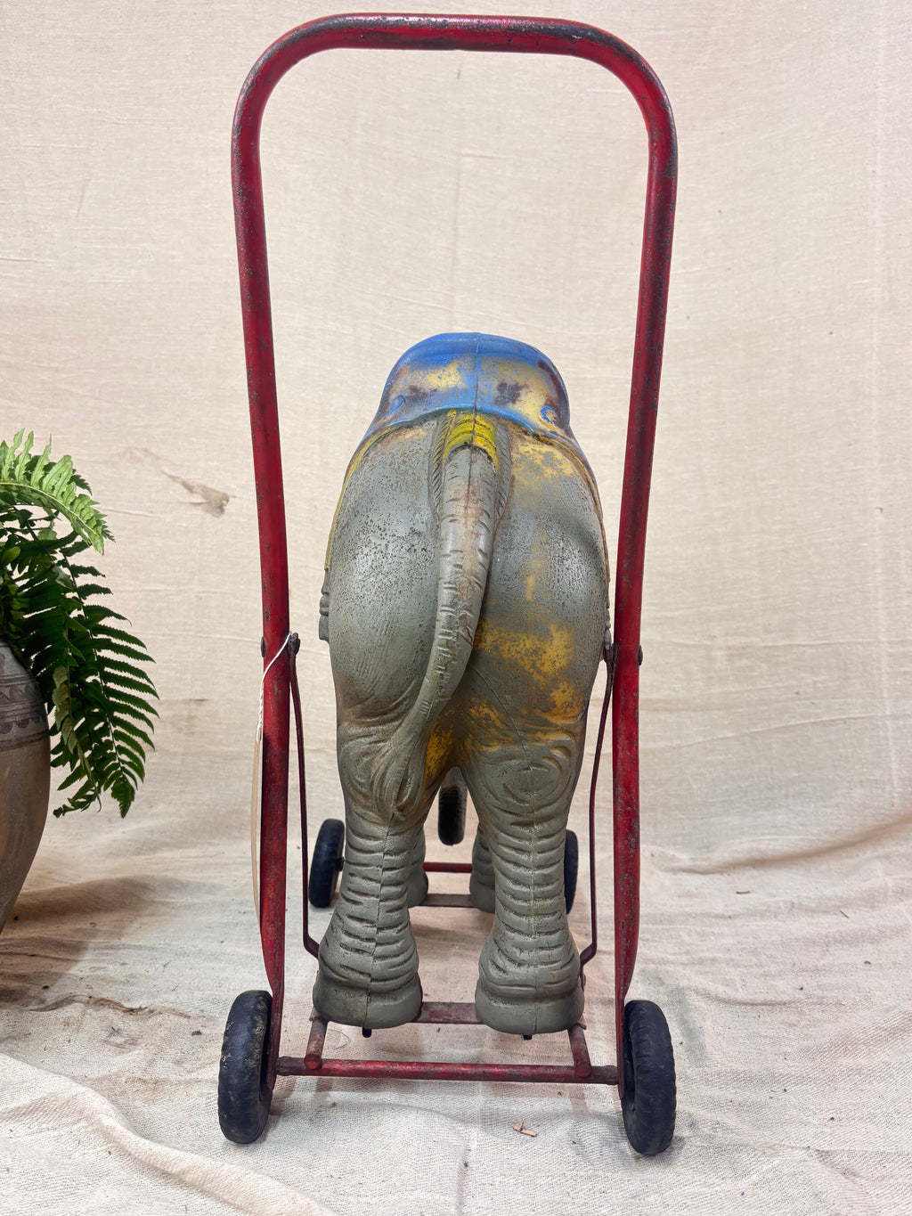 Vintage 1950s Tri-ang push-along elephant toy with hard plastic body, blue saddle, and red metal chassis with rubber wheels with a plant on a cream background