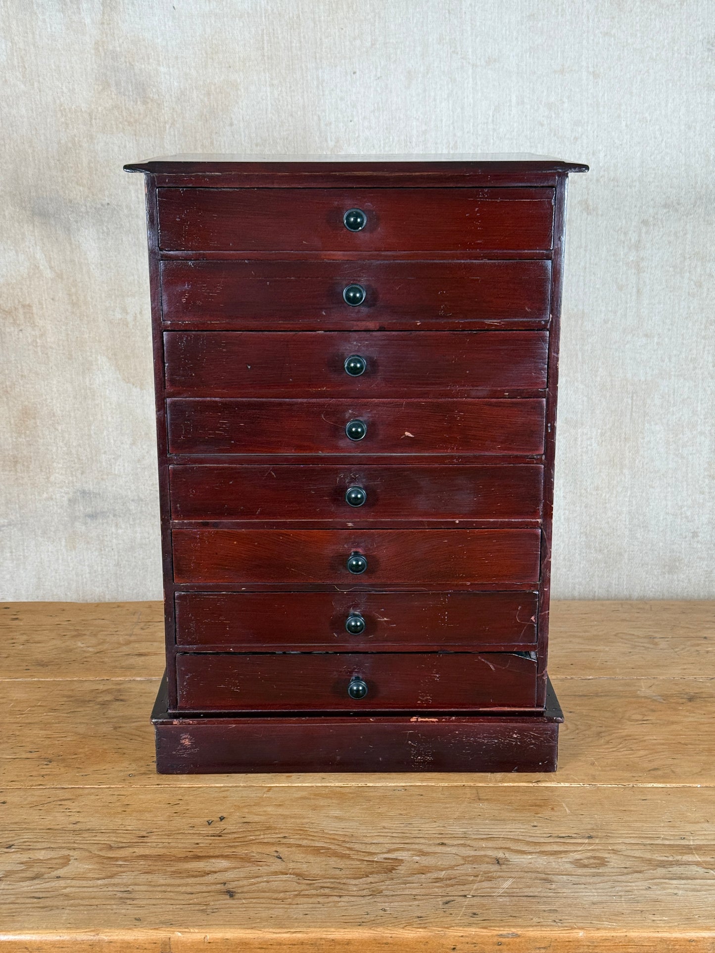 A vintage dark reddish-brown wooden chest of drawers with eight drawers and small round black knobs, standing on a light wood table against a textured beige wall.