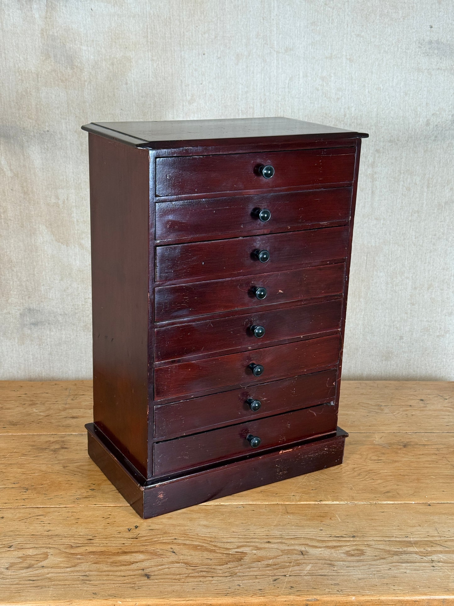 A vintage dark reddish-brown wooden chest of drawers with seven drawers and small round black knobs, standing on a wooden surface against a textured beige wall.