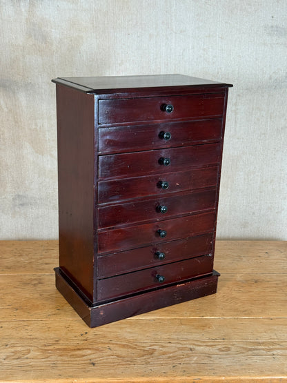 A vintage dark reddish-brown wooden chest of drawers with seven drawers and small round black knobs, standing on a wooden surface against a textured beige wall.