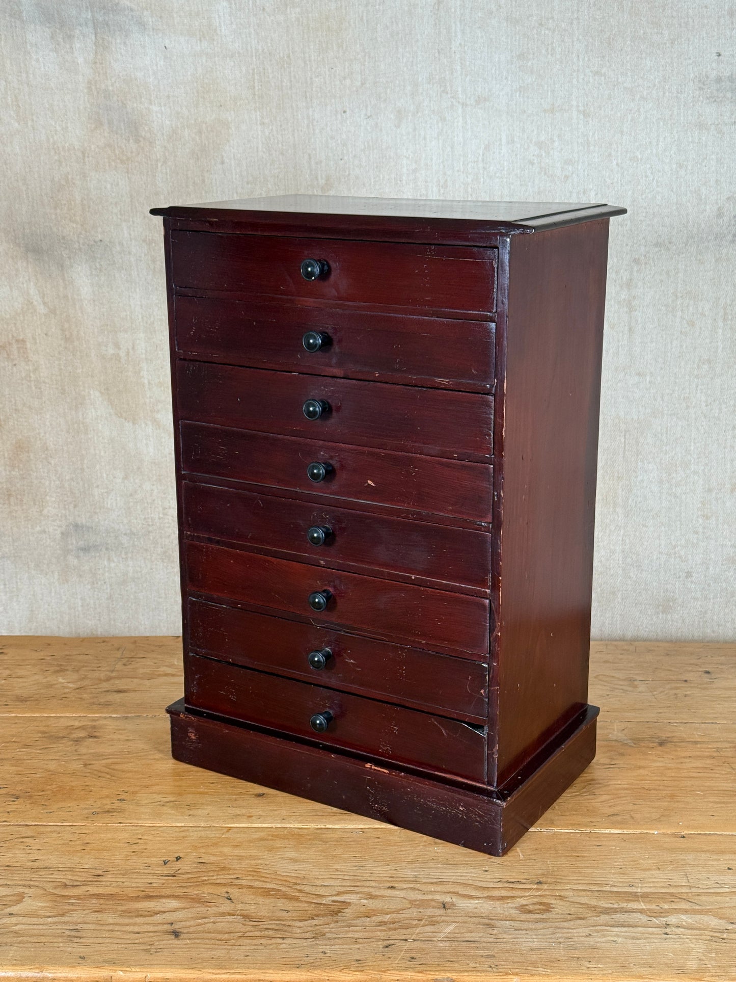 A vintage dark reddish-brown wooden chest of drawers with seven drawers and small round black knobs, set against a neutral textured wall and wooden floor.