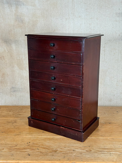 A vintage dark reddish-brown wooden chest of drawers with seven drawers and small round black knobs, set against a neutral textured wall and wooden floor.