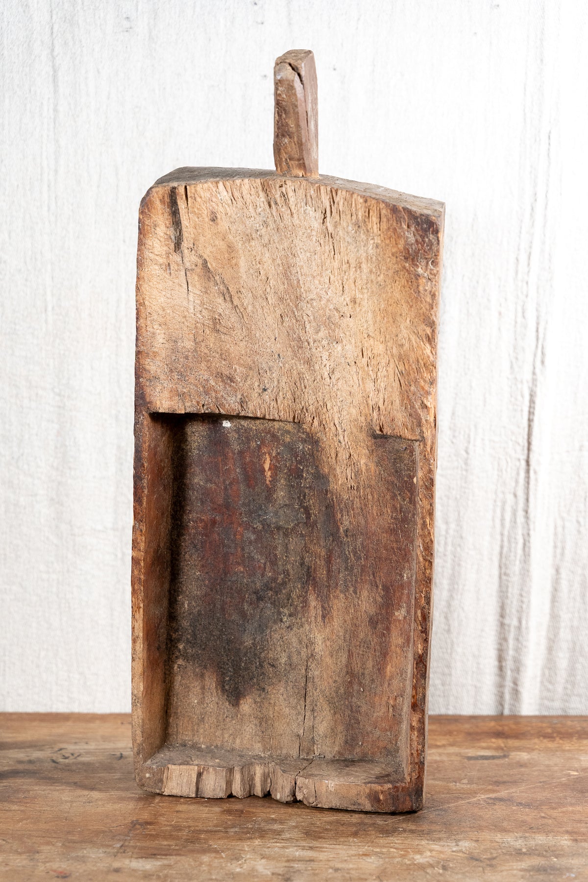 A rustic, hand-carved wooden box or container with a loop handle on top, standing on a wooden surface against a white textured background.