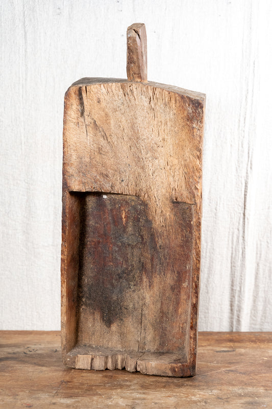 A rustic, hand-carved wooden box or container with a loop handle on top, standing on a wooden surface against a white textured background.
