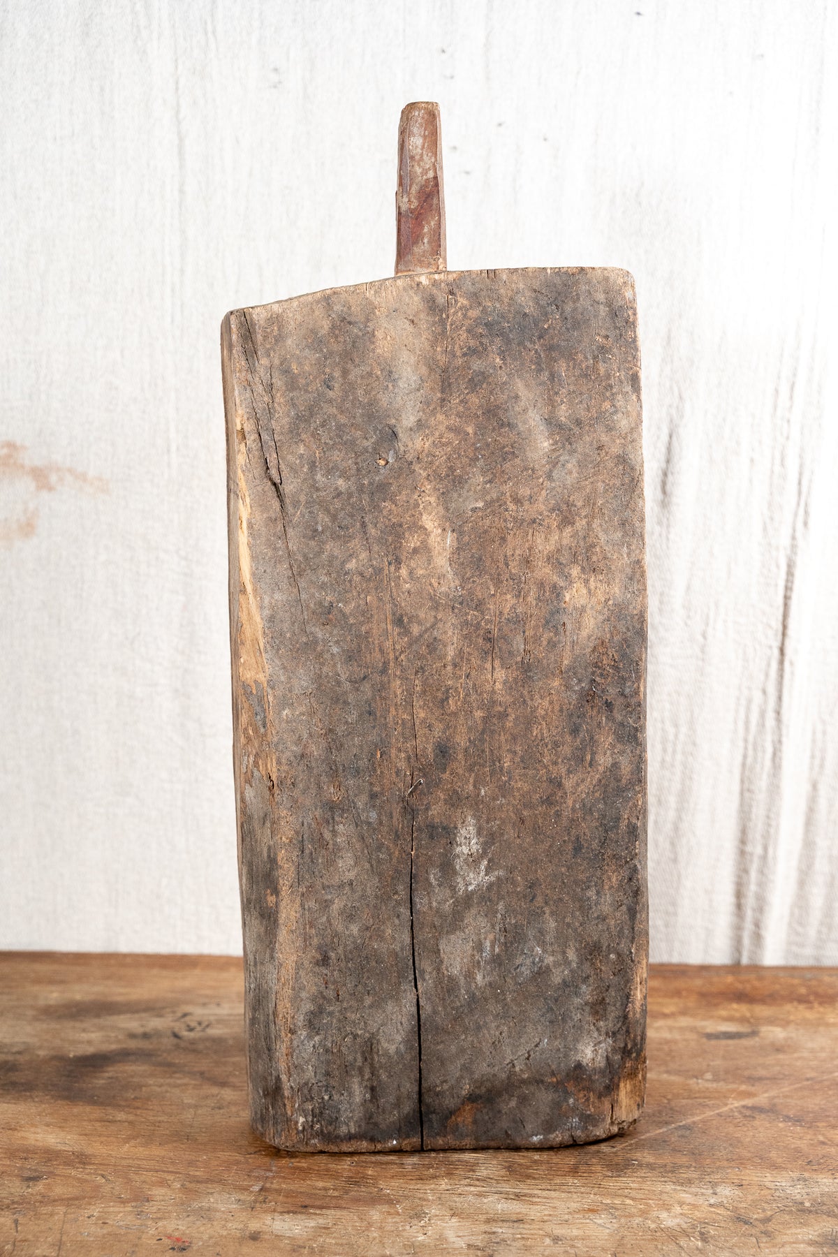 A rustic, hand-hewn rectangular wooden cutting board with a single peg handle, standing upright on a wooden surface against a white wood plank background.