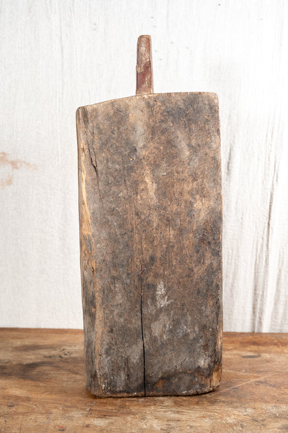 A rustic, hand-hewn rectangular wooden cutting board with a single peg handle, standing upright on a wooden surface against a white wood plank background.