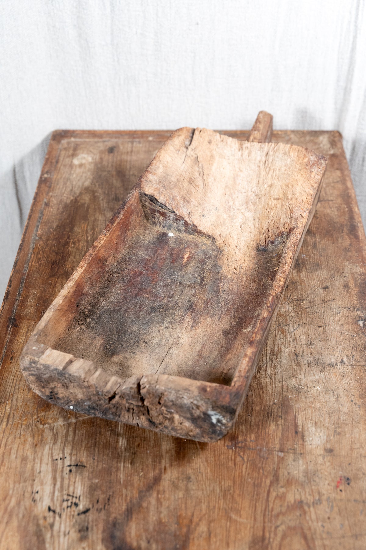 A rustic, hand-carved wooden scoop or trough with a long handle, resting on a weathered wooden table against a white fabric backdrop.