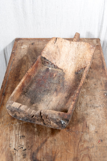 A rustic, hand-carved wooden scoop or trough with a long handle, resting on a weathered wooden table against a white fabric backdrop.