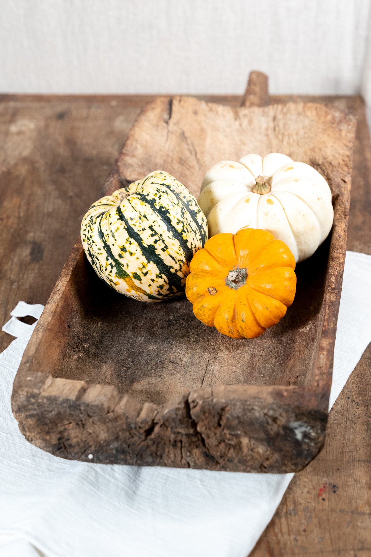 Three decorative pumpkins and gourds arranged in a rustic wooden tray on a wooden table with a white cloth underneath.