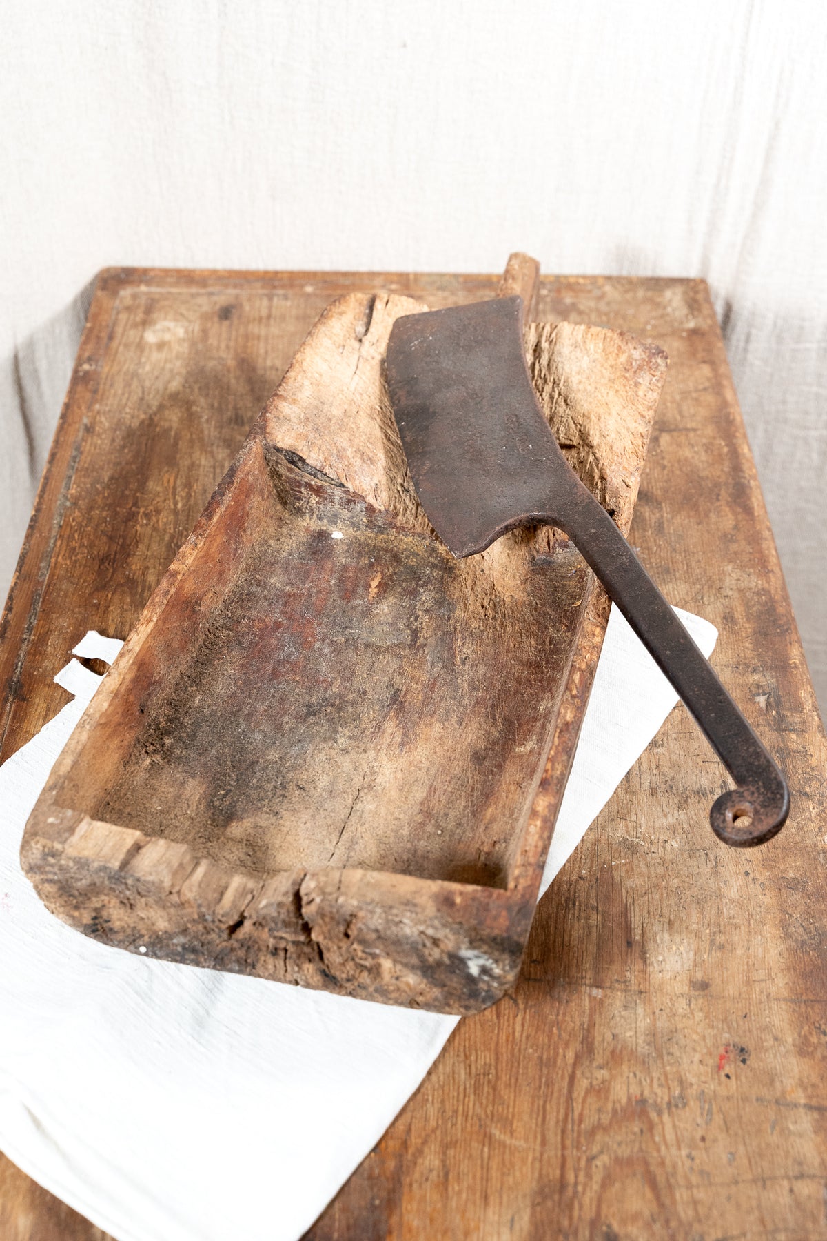 A rustic wooden trough or mold with a dark metal spatula resting inside it, placed on a white cloth over a weathered wooden surface.