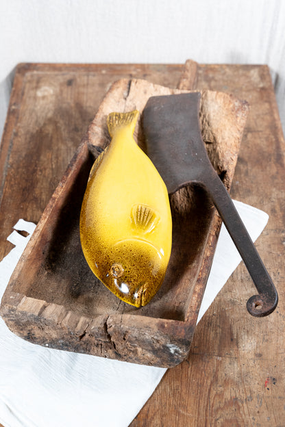 A glossy yellow glass fish sculpture sits in a rustic wooden tray with a dark metal butcher-style knife prop, set on a weathered wooden board with a white cloth underneath.