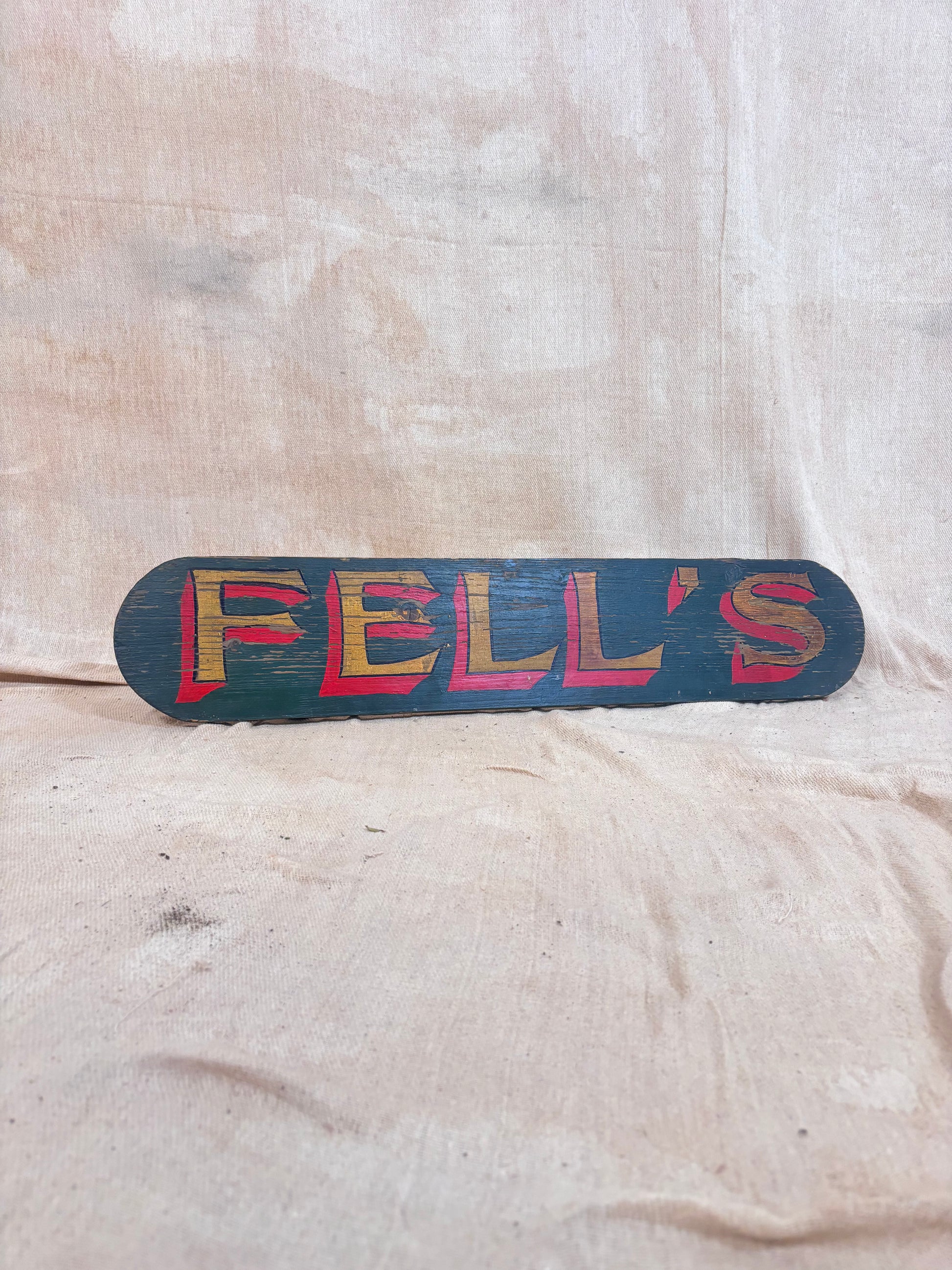A rustic, hand-painted wooden sign reading 'FELL'S' in red and yellow lettering on a dark teal background, displayed against a draped beige fabric backdrop.