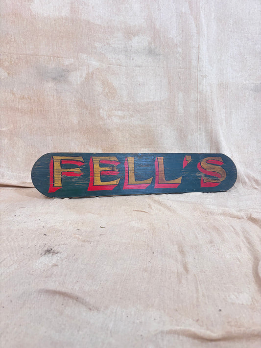 A rustic, hand-painted wooden sign reading 'FELL'S' in red and yellow lettering on a dark teal background, displayed against a draped beige fabric backdrop.