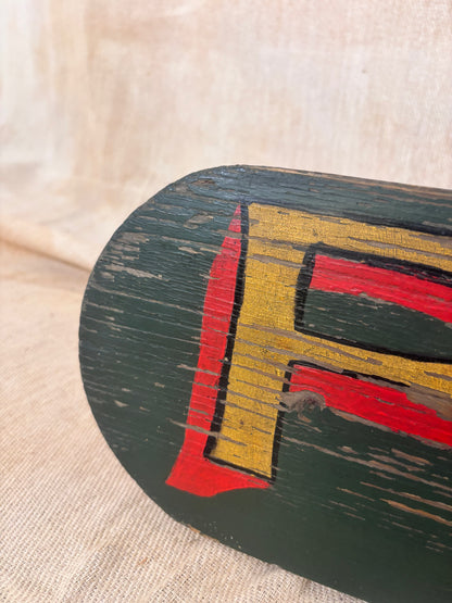 A close-up of a dark green wooden sign featuring a large distressed yellow and red painted letter 'F' on a beige fabric background.