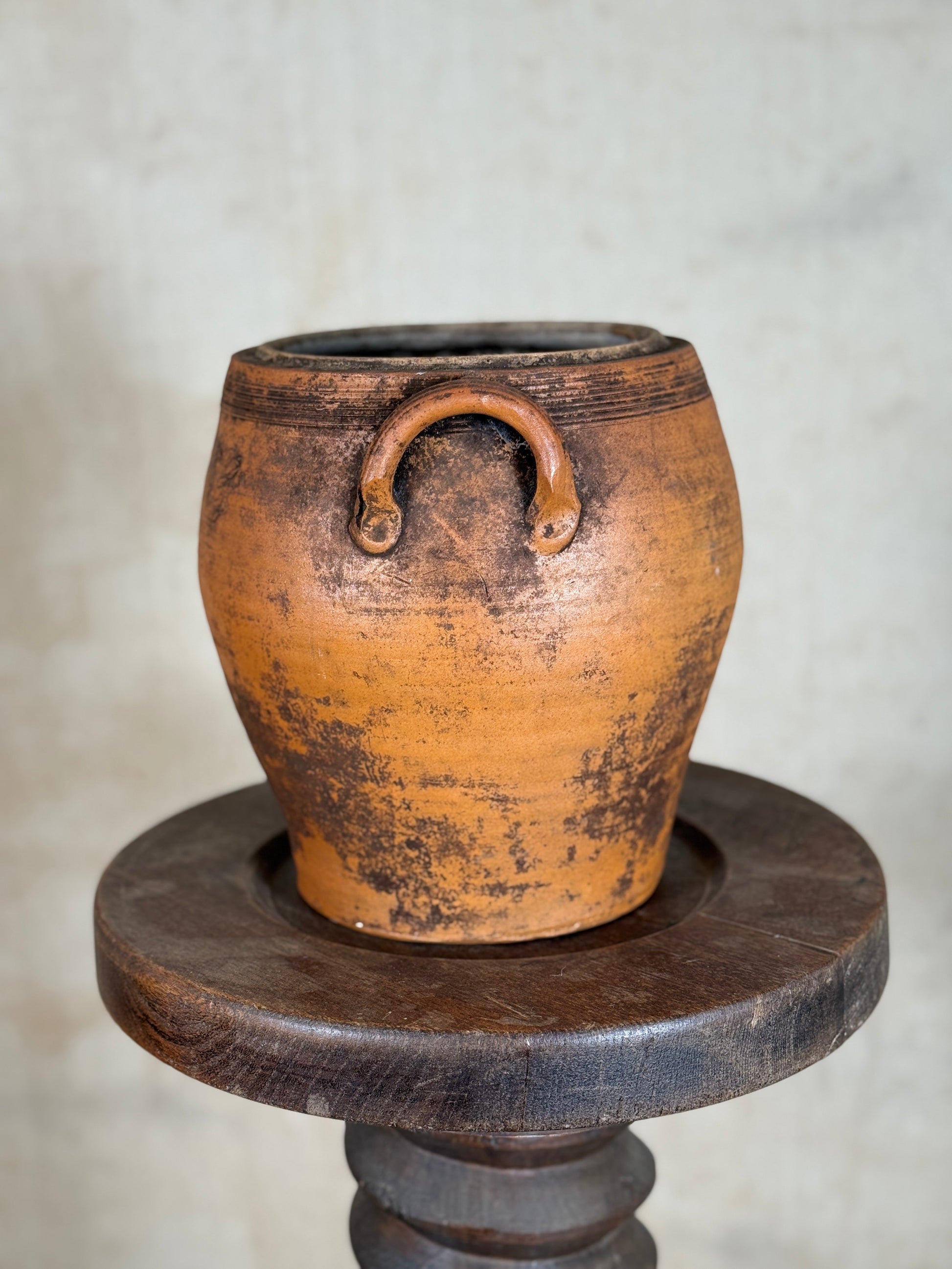 An antique terracotta clay pot with a rustic brown handle, displayed on a dark wooden pedestal against a neutral textured background.